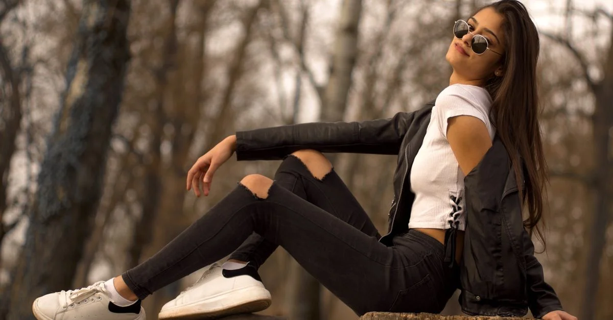 Jeune femme élégante en lunettes de soleil et veste en cuir posant en plein air sur une souche d'arbre.