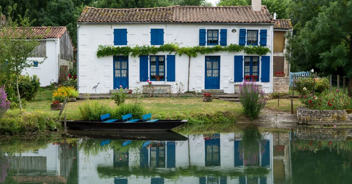 Charmante maison blanche avec volets bleus au bord d'un canal paisible, entourée de verdure.