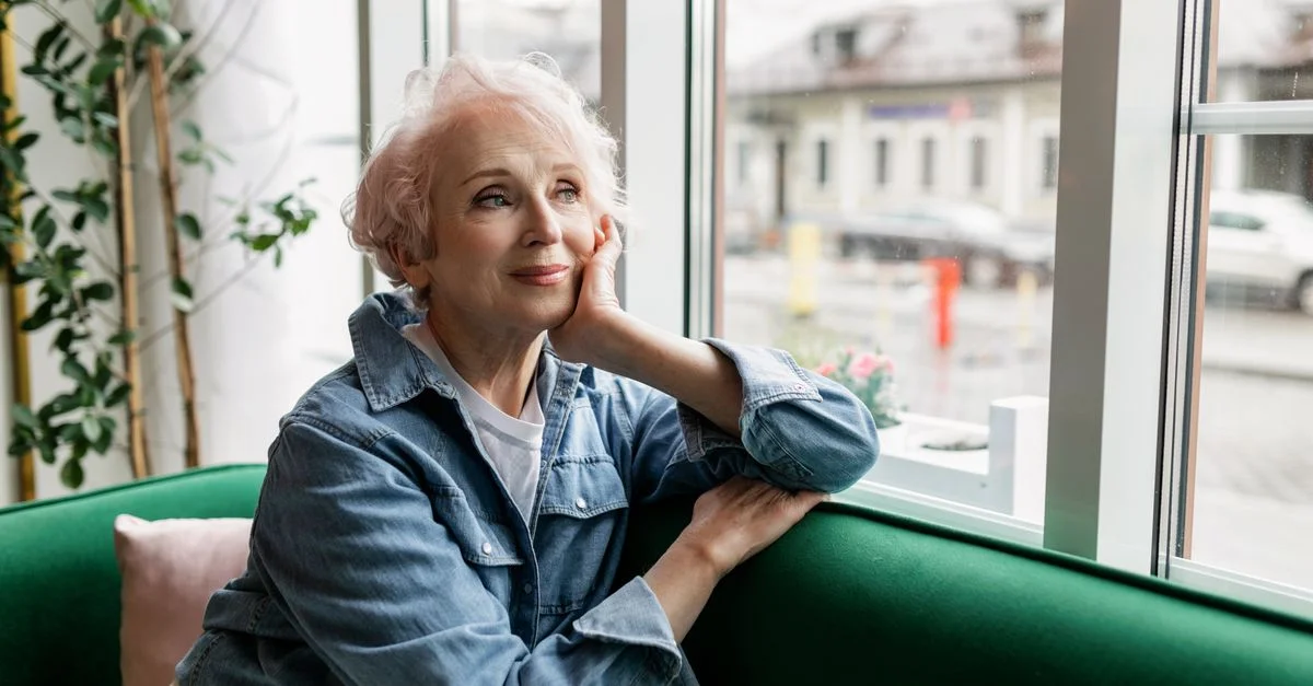 Femme senior gracieuse en veste en jean assise près d'une fenêtre, réfléchissant tranquillement à l'intérieur.