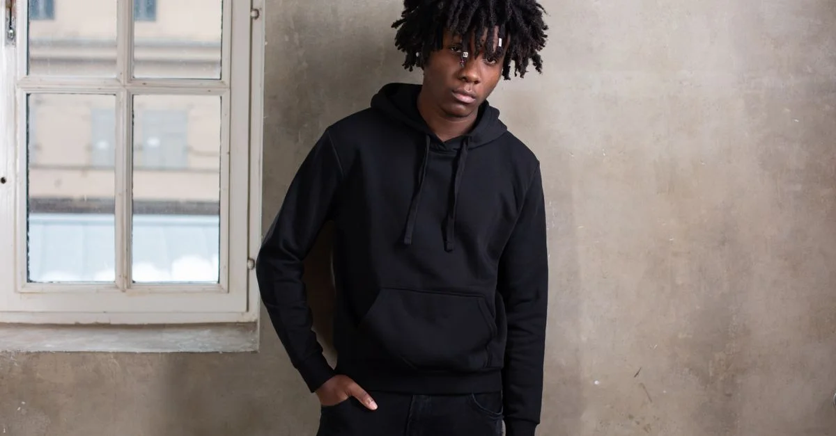 Un jeune homme aux dreadlocks dans un hoodie noir se tient avec confiance à l'intérieur près d'une fenêtre.