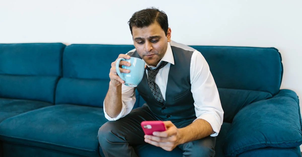 Un homme en costume se détend sur un canapé bleu, buvant du café et utilisant un smartphone.