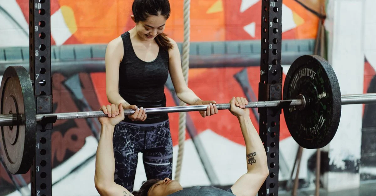 Femme aidant un homme lors d'un exercice de développé couché dans une salle de sport colorée.