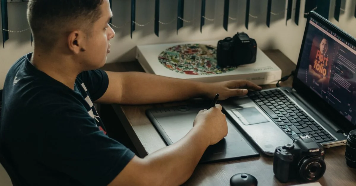 Homme retouchant des photos sur un ordinateur portable à l'aide d'une tablette graphique, situé dans un espace de travail intérieur avec du matériel photo.