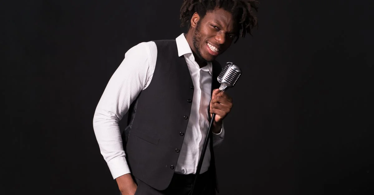 Homme souriant en gilet tenant un microphone vintage sur un fond sombre, dégageant du charisme.